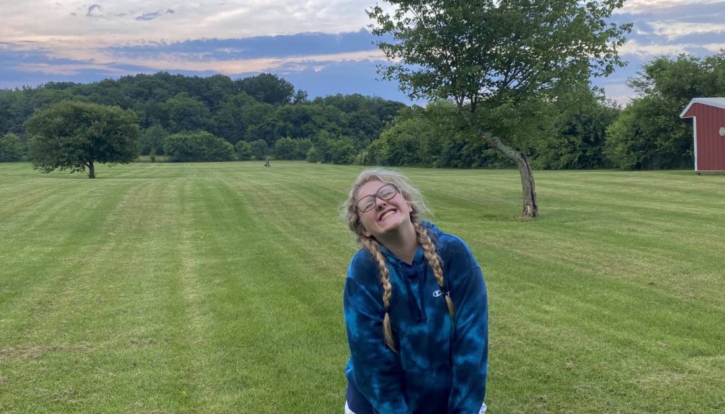 A smiling young woman with braided blonde hair and glasses wearing a blue tie-dye hoodie in a vast grassy field.