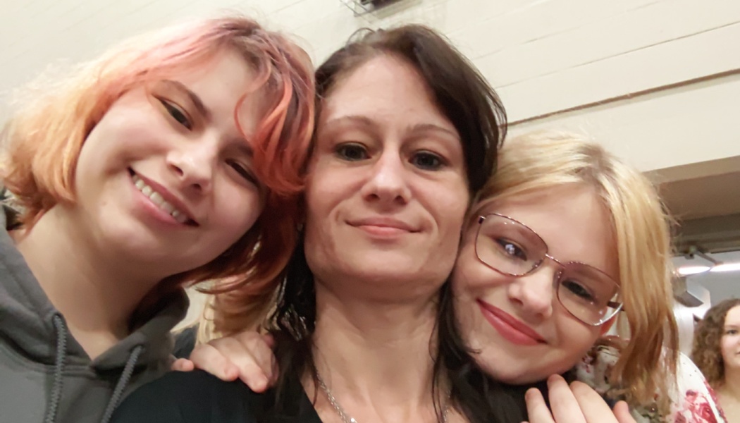 A smiling woman poses for a close selfie with two young girls leaning against her.