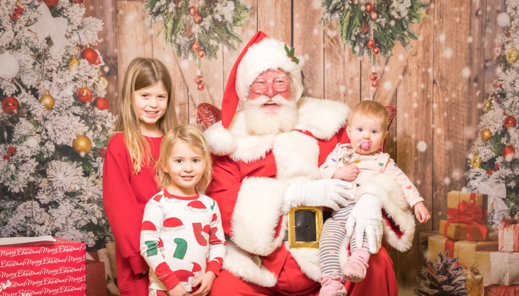 Santa Claus holding a baby while two young girls smile beside him in front of a festive Christmas backdrop.