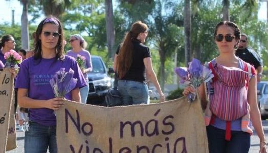 Women march holding purple flowers and a banner reading "No más violencia," one carrying a baby.