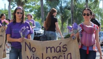 Women march holding purple flowers and a banner reading "No más violencia," one carrying a baby.