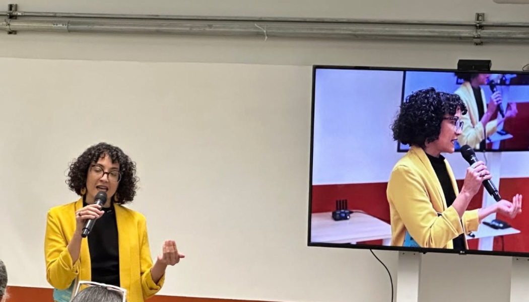 A woman in a yellow blazer speaks into a microphone next to a screen displaying a live feed of her speaking.