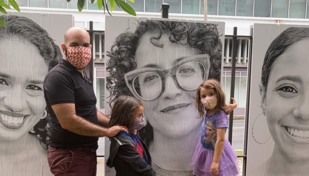 A masked man and two young children stand outdoors in front of large black-and-white portrait posters.