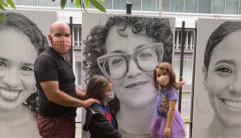 A masked man and two young children stand outdoors in front of large black-and-white portrait posters.