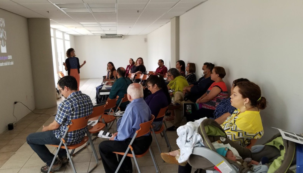 A woman presents to a seated audience of community members in a bright meeting room.