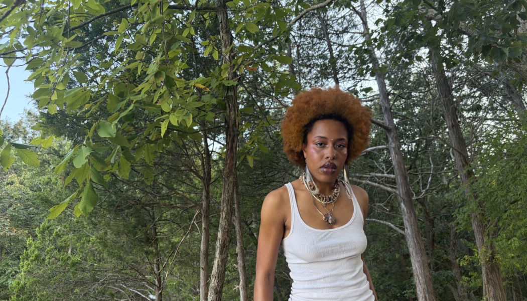 A Black woman with a reddish afro and shell necklaces stands outdoors in a lush, wooded forest.
