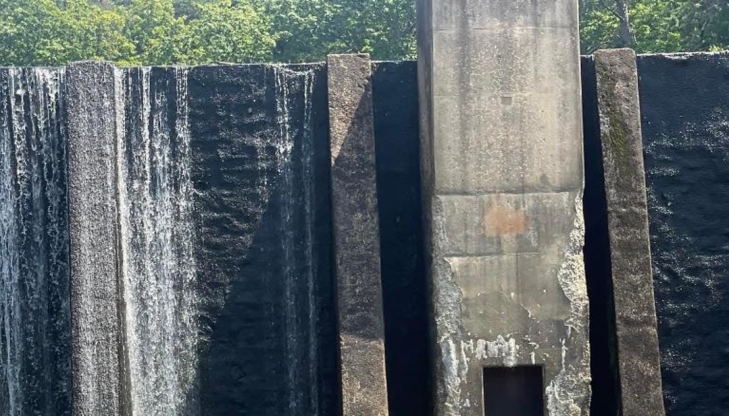 Water cascading over a tall, weathered concrete dam with green forest at the top.