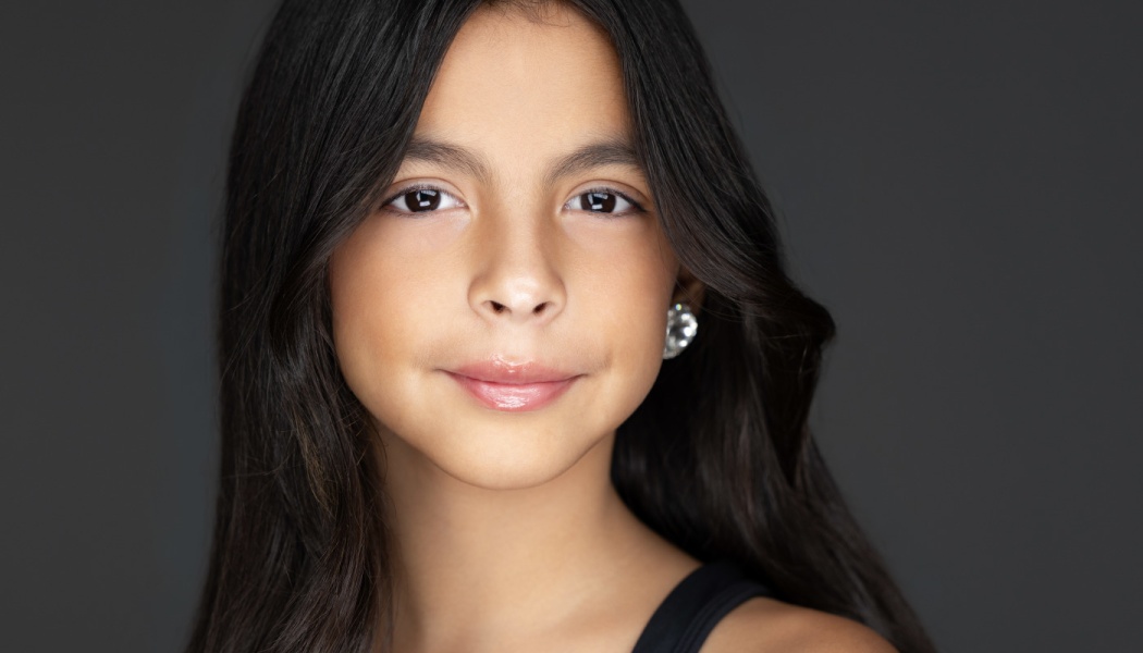 Close-up headshot of a young girl with long dark hair, brown eyes, and a gentle smile against a dark background.
