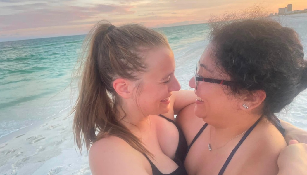 Two women embrace and smile lovingly at each other on a sandy beach at sunset with the ocean in the background.