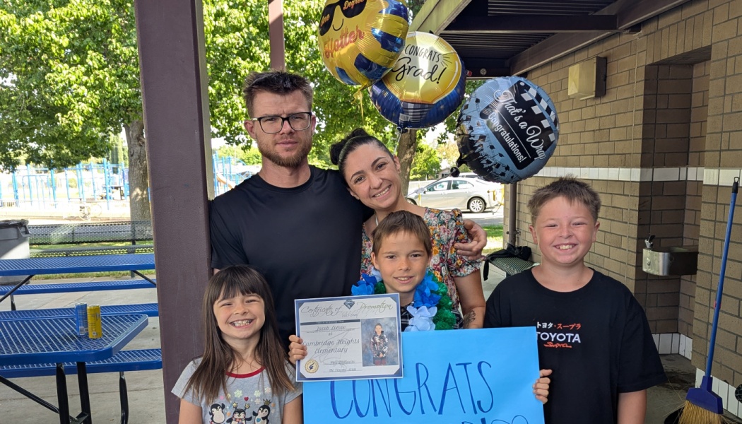 A happy family of five posing outdoors with graduation balloons and an elementary school promotion certificate.