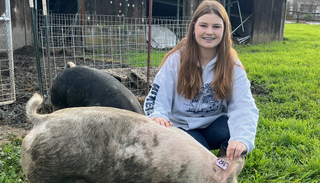 A smiling girl in a gray hoodie kneels in grass while petting a large farm pig near a barn.