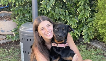 A smiling young woman hugs a small black and tan dog making a silly side-eye expression outdoors.
