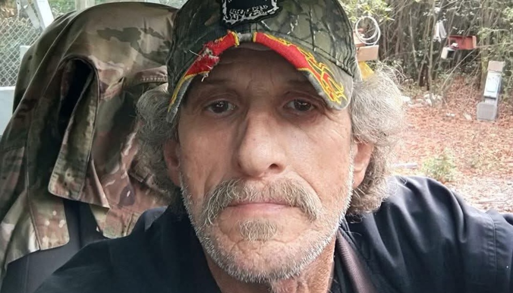 A man with wavy gray hair, a mustache, and a worn camo baseball cap looking at the camera.