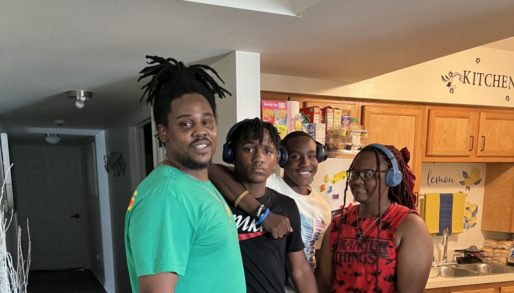 A Black father and his three teenage children pose closely together in their kitchen.