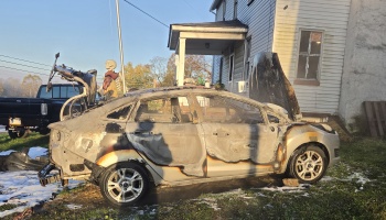 A heavily fire-damaged silver sedan with charred, melted paint parked outside a residential house.