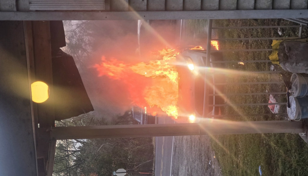 A car engulfed in bright orange flames and heavy smoke, viewed from a porch.