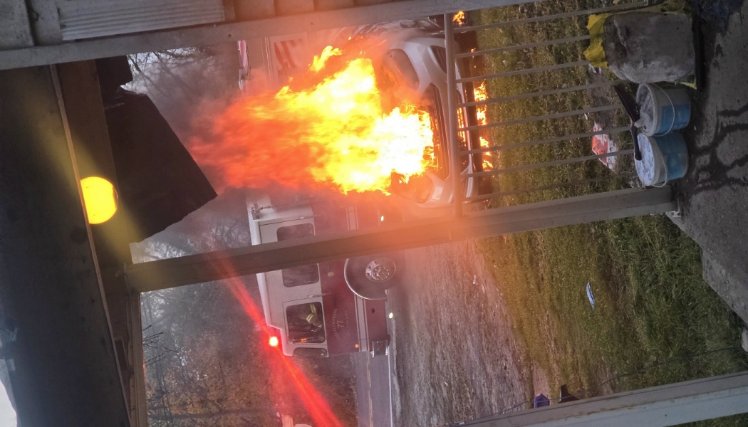 A car fully engulfed in bright orange flames outside a house, with a fire truck parked nearby on the street.