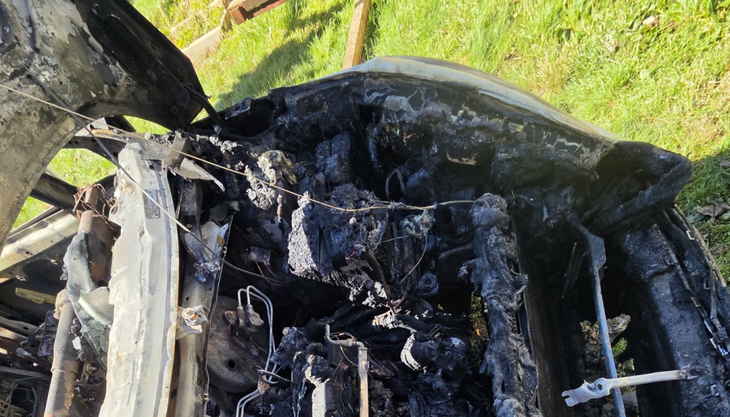 Severely burned car engine bay with charred wires, melted plastic, and extensive fire damage.