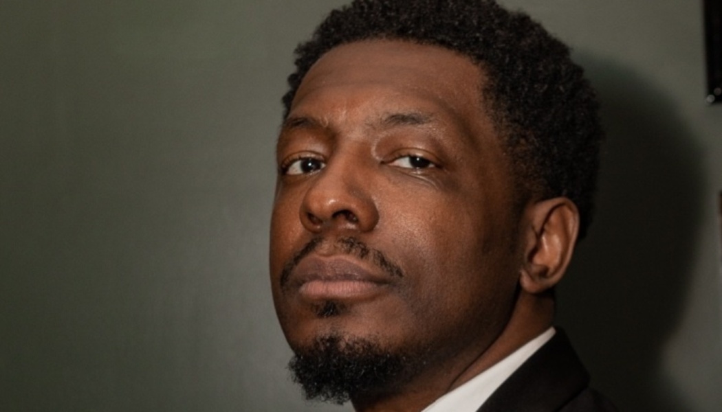 Close-up portrait of a Black man with a goatee, wearing a dark suit jacket and white shirt.