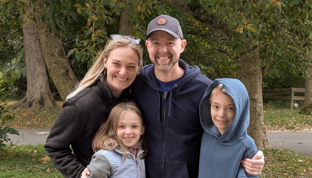 A happy family of four—a mother, father, daughter, and son—smiling together outdoors in a park.