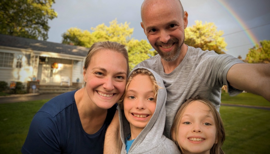 Smiling family of four taking a selfie outdoors in front of their home with a rainbow in the background.