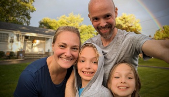 Smiling family of four taking a selfie outdoors in front of their home with a rainbow in the background.