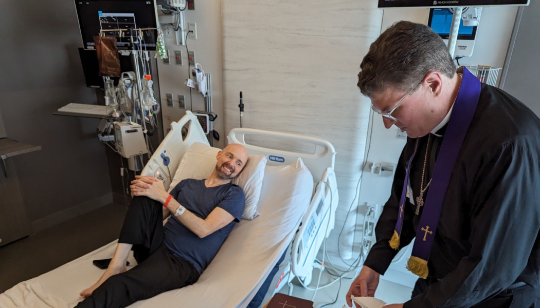 A smiling man in a hospital bed looks up at a priest wearing a purple stole standing at his bedside.