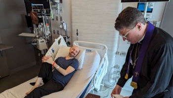 A smiling man in a hospital bed looks up at a priest wearing a purple stole standing at his bedside.