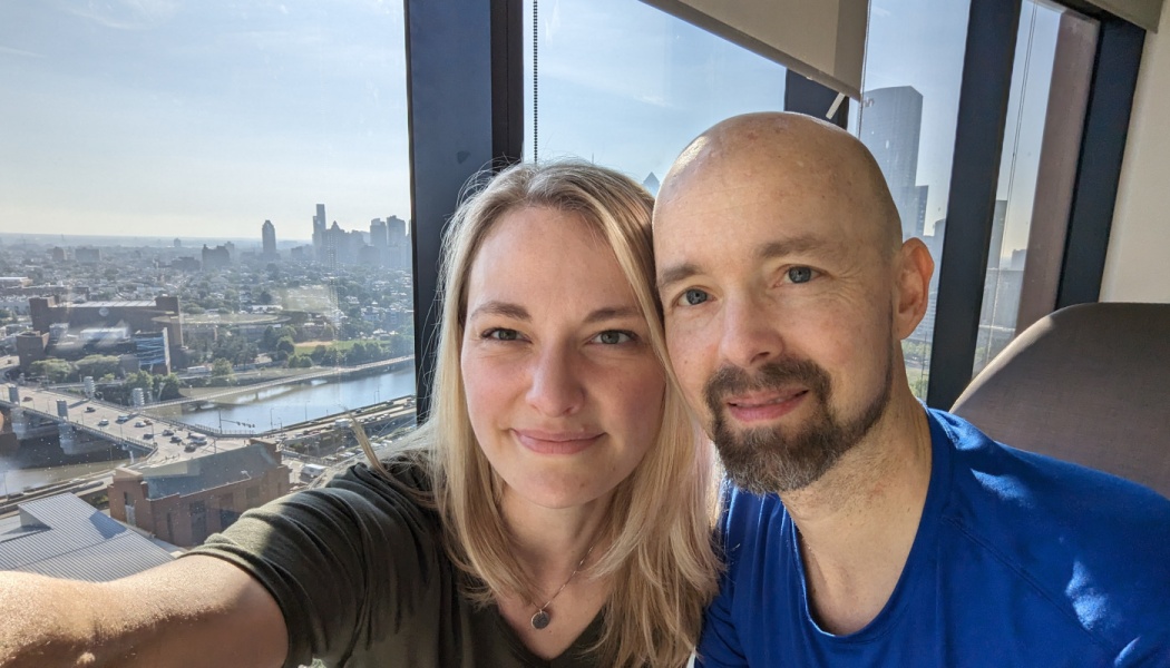 A smiling blonde woman and a bald man take a selfie in front of a high-rise window overlooking a city skyline.