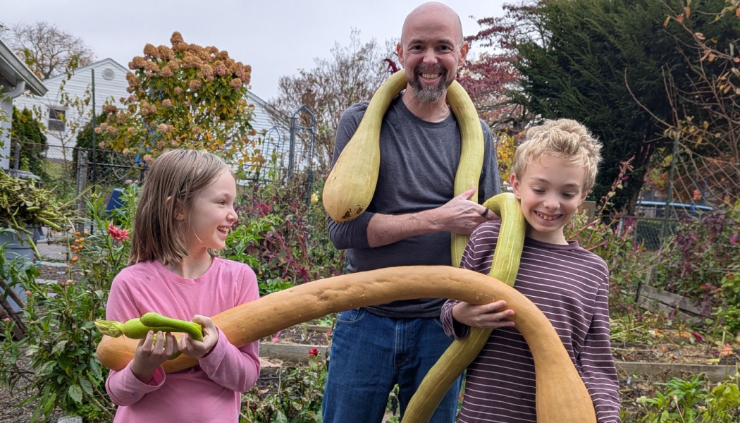 A smiling father and two children in a garden pose with giant, long, curved squash draped around them.