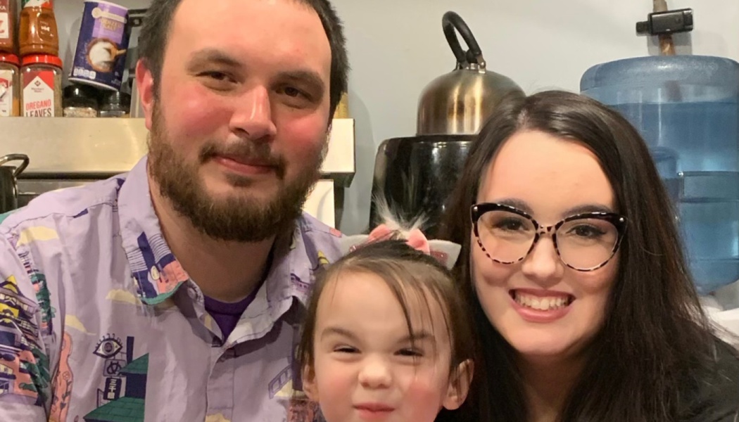 A smiling man with a beard, a woman with glasses, and a young child pose close together indoors.
