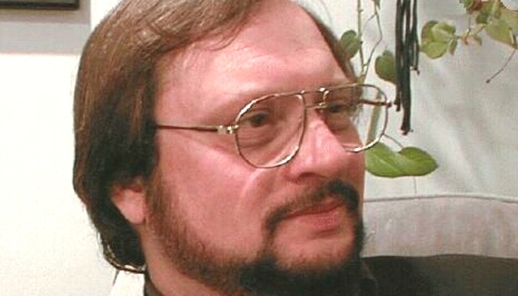 Close-up of a man with brown hair, wire-rimmed glasses, and a beard looking thoughtfully off-camera.