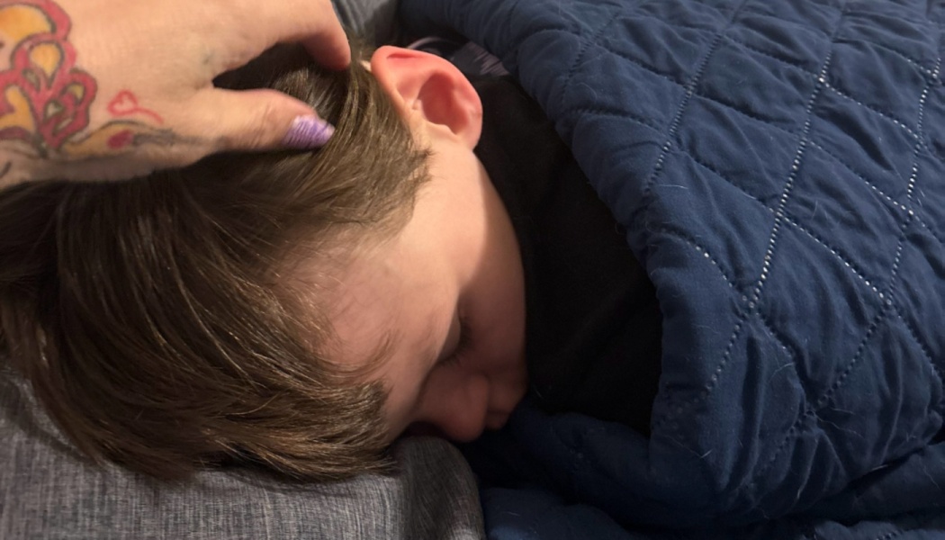A tattooed hand gently strokes the hair of a sleeping child tucked under a blue quilted blanket.