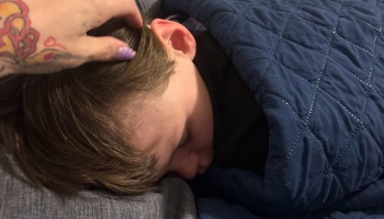 A tattooed hand gently strokes the hair of a sleeping child tucked under a blue quilted blanket.