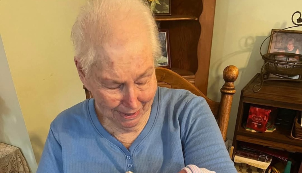 An elderly woman with white hair wearing a blue shirt looks down tenderly while holding a newborn baby.