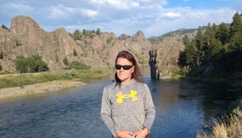 A woman wearing sunglasses and a grey hoodie stands before a river flowing through a dramatic, rocky canyon.