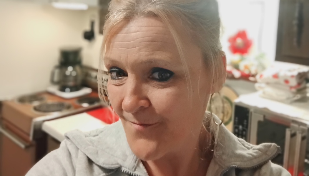 Blonde woman wearing a gray sweater smirking slightly in her kitchen.