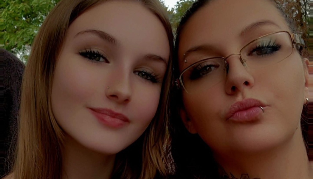 Close-up selfie of two young women posing together, one smiling softly and the other wearing glasses.