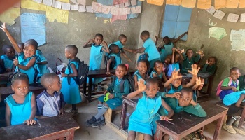 Young African students in blue uniforms smiling and waving from wooden desks in a crowded classroom.