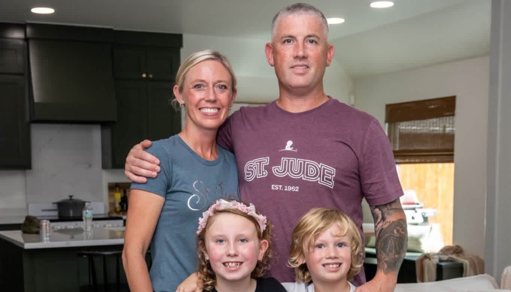 A smiling family of four—a mother, father, and two young children—pose together in their kitchen.