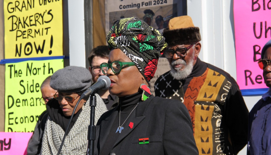 A woman in a colorful headwrap speaks into a microphone at a community rally, flanked by supporters and signs.