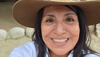 Close-up of a smiling woman with long dark hair wearing a wide-brimmed brown hat and a white shirt.