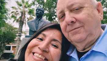 A smiling woman and an older man take a close-up selfie outdoors with a bronze bust statue behind them.