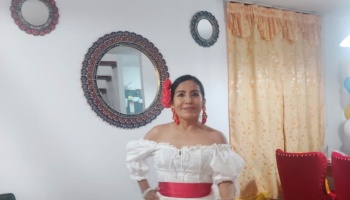 A smiling woman wearing a traditional white off-the-shoulder dress with a red sash and a red flower in her hair.
