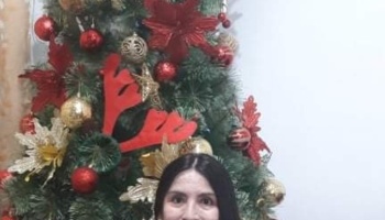 A dark-haired woman standing in front of a Christmas tree decorated with red and gold ornaments.