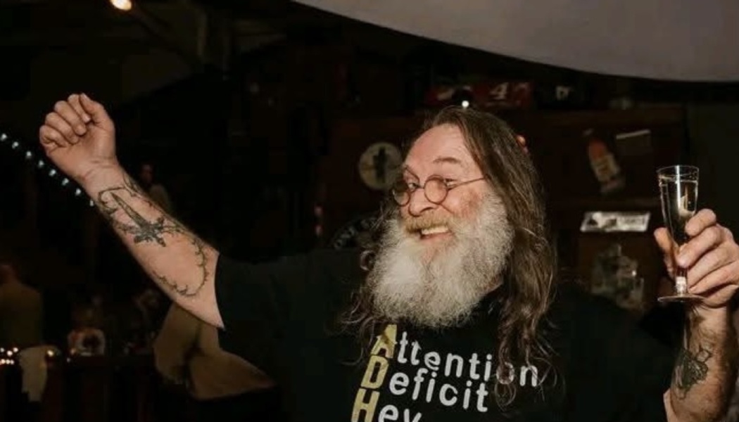 Smiling man with a long white beard raising his tattooed arm and holding a champagne flute in celebration.