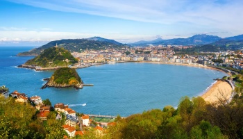 Panoramic view of a crescent-shaped turquoise bay with a sandy beach, a small island, and a coastal city.