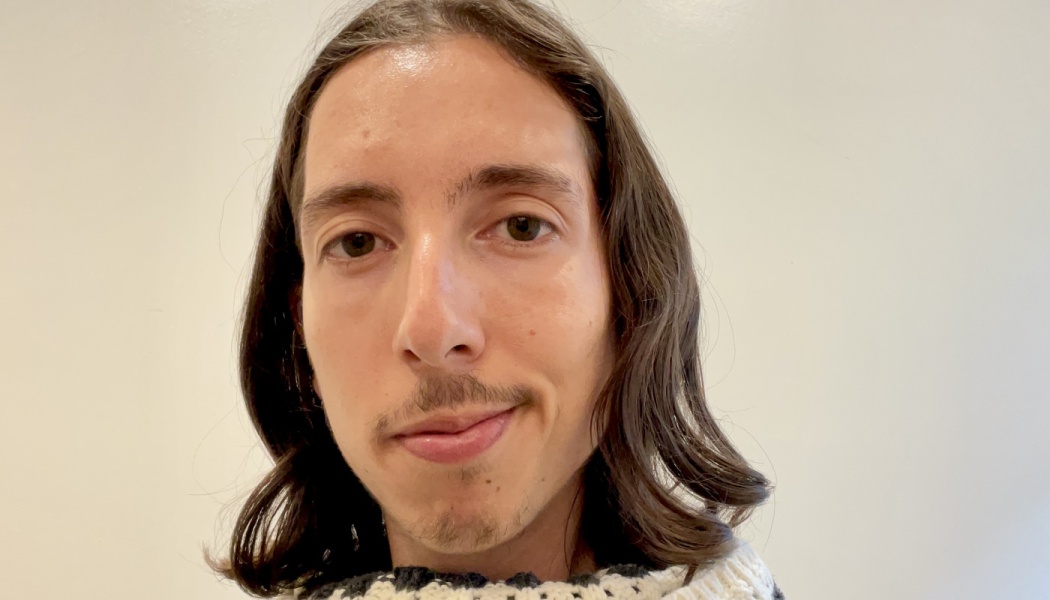Young person with long, wavy brown hair and a slight mustache looking at the camera with a gentle smile.
