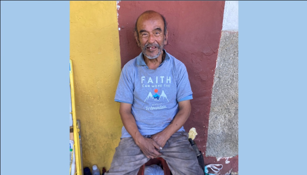 An older man with a warm smile wearing a blue t-shirt, sitting in front of a colorful yellow and red wall.