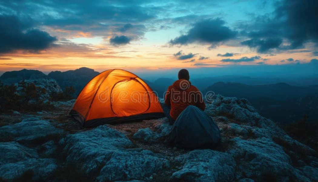 A camper sits on a rocky mountain peak next to a glowing orange tent at dusk, overlooking distant peaks.